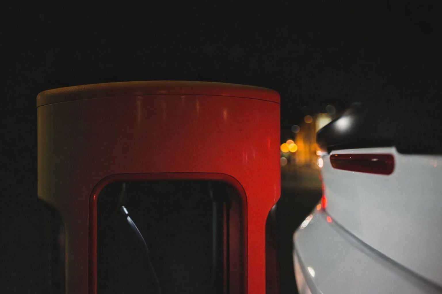 Röd Zaptec Pro-laddstation intill en vit elbil nattetid, med suddiga lampor i bakgrunden.